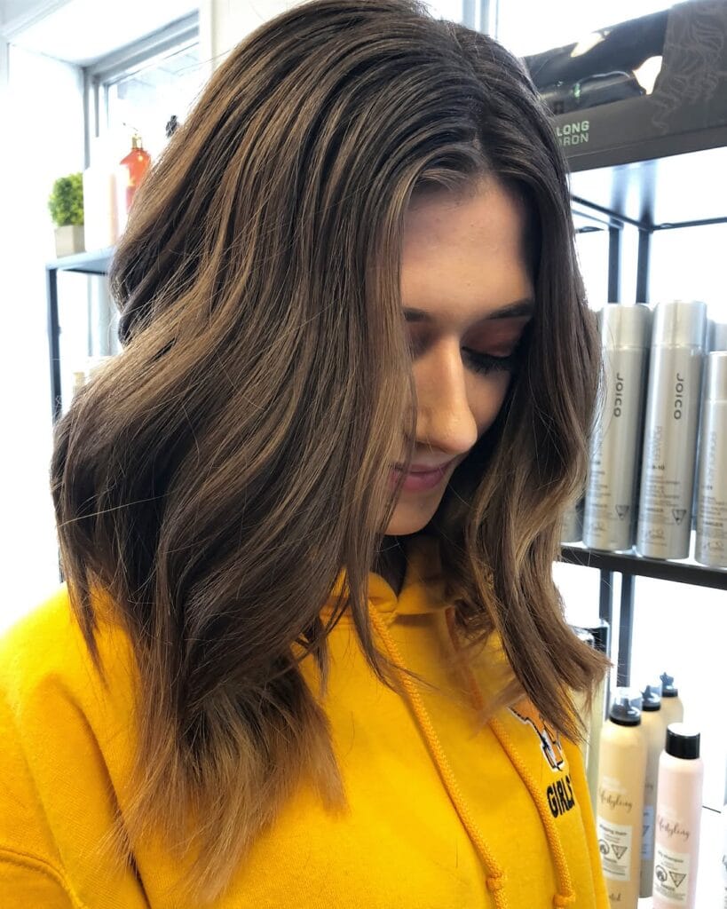 A woman with shoulder length dark brown hair featuring subtle blonde balayage styled in loose relaxed waves.