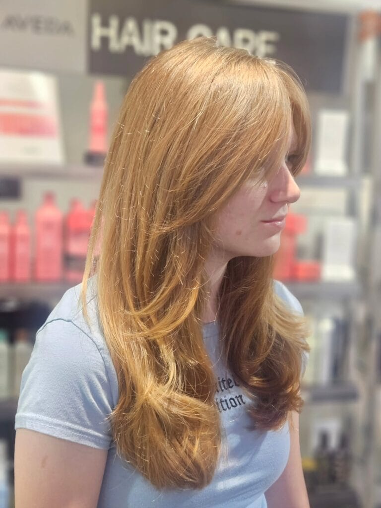 A woman viewed from the side with a voluminous long layered copper haircut featuring swooping face-framing fringe.