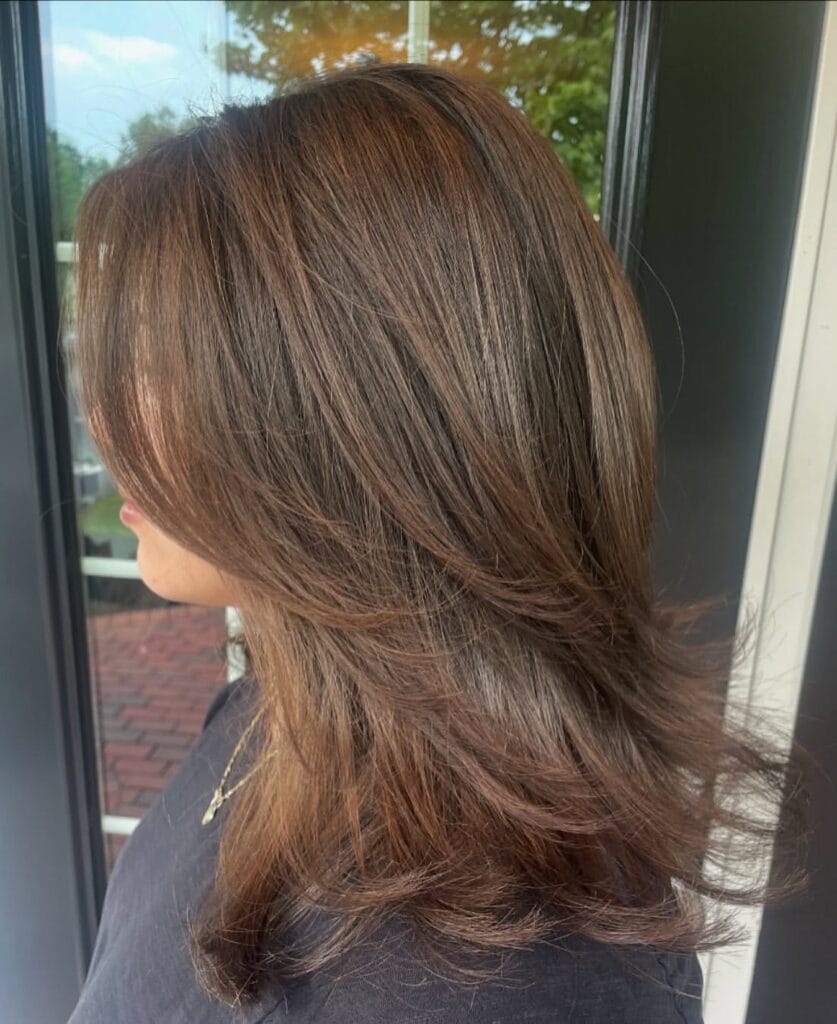 A woman viewed from the side with medium-length layered brown hair styled with voluminous ends.