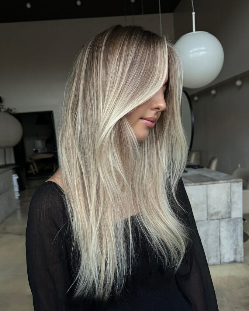 A woman with long straight hair featuring a bright champagne blonde balayage and soft face-framing layers.