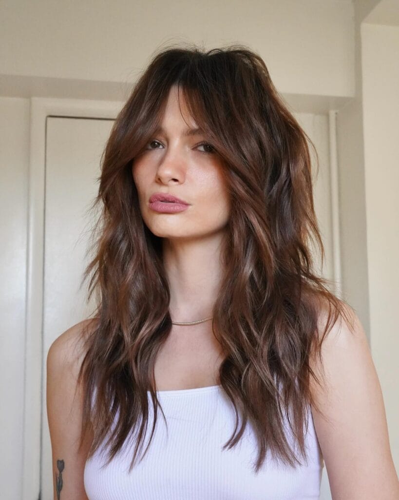 A woman with long shaggy brunette hair featuring heavy layers and face-framing curtain bangs.