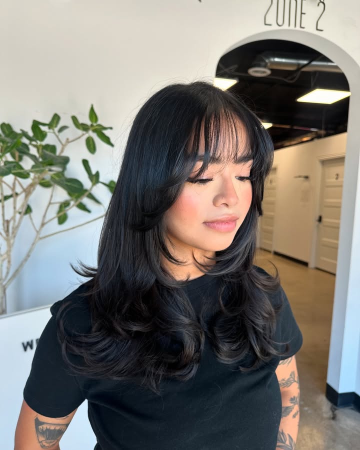 Long layered dark hair with full soft bangs and curled ends for volume.