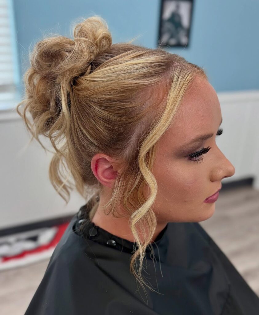 A side profile photograph of a young woman featuring a voluminous high bun in multi-toned blonde hair with a wavy piece framing her face.