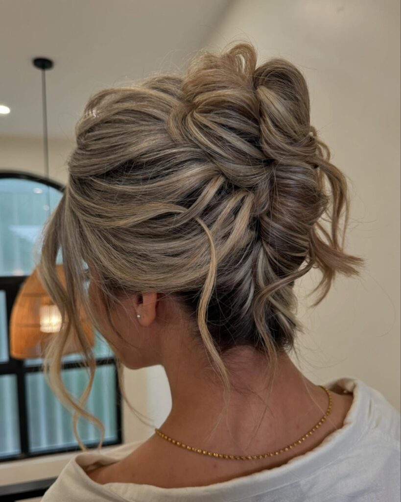 A woman with sandy blonde highlighted hair styled in a textured and voluminous wrap updo with loose wispy strands.