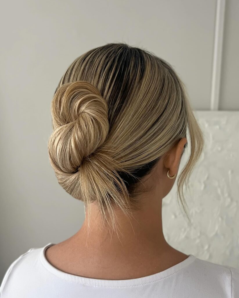 A back view of a woman with blonde highlighted hair styled in a sleek vertical tucked bun with a soft strand framing her face.