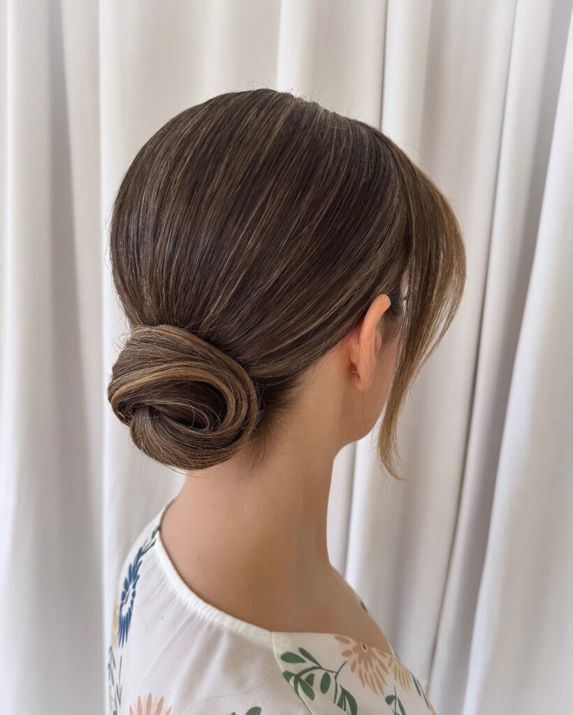 A side profile of a woman wearing a sleek low bun with blonde highlights and soft side bangs against a white curtain background.
