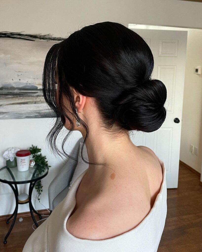 A woman with dark black hair styled in a large smooth low bun with long wavy face framing tendrils.