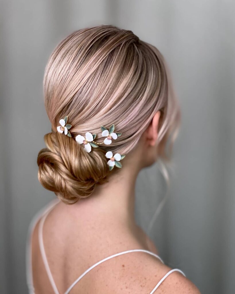 A back view of a woman with rose gold hair styled in a low bun decorated with small white flower hair clips.