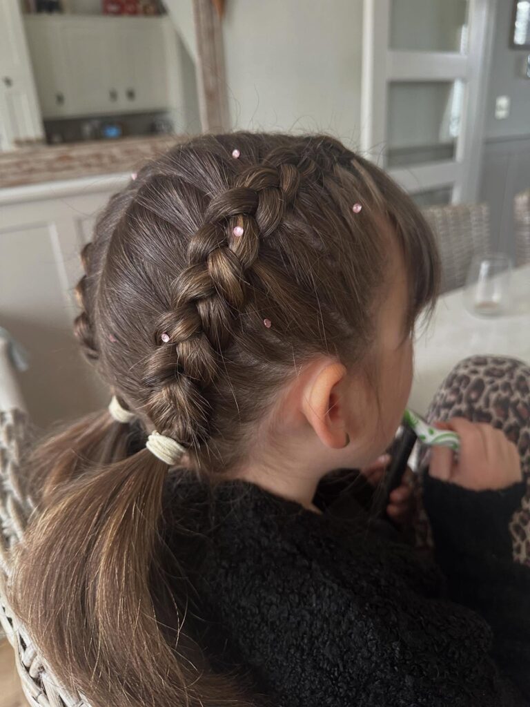 A young girl seen from the side wearing thick double Dutch braids accented with small pink rhinestones and finished with low pigtails.