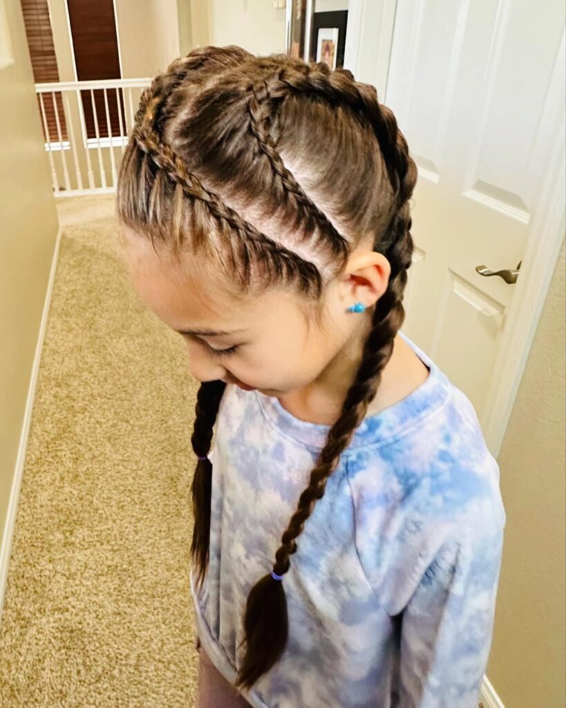 A young girl looking down, showing her brunette double Dutch braids with additional smaller accent braids along the side of her head.