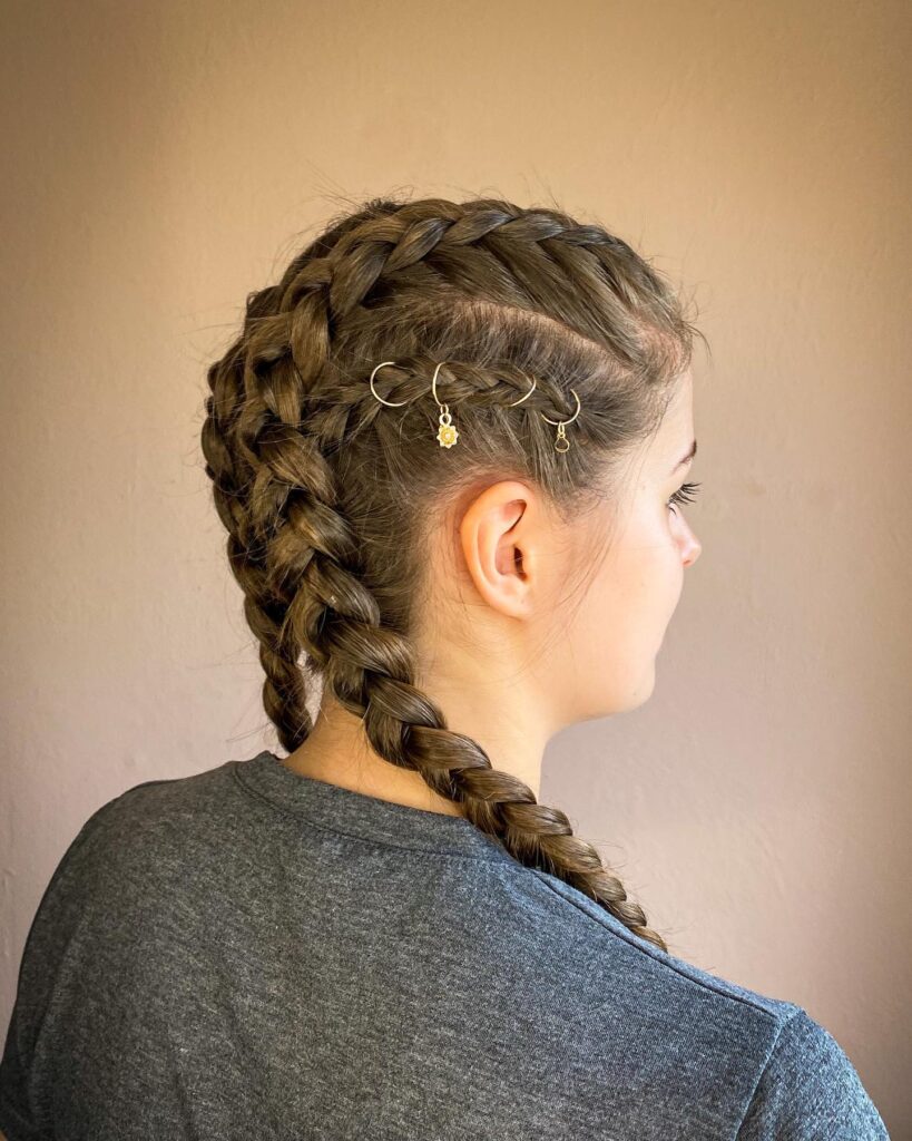 A woman seen from the side showing brunette double Dutch braids accented with small gold hair rings and a sun charm.