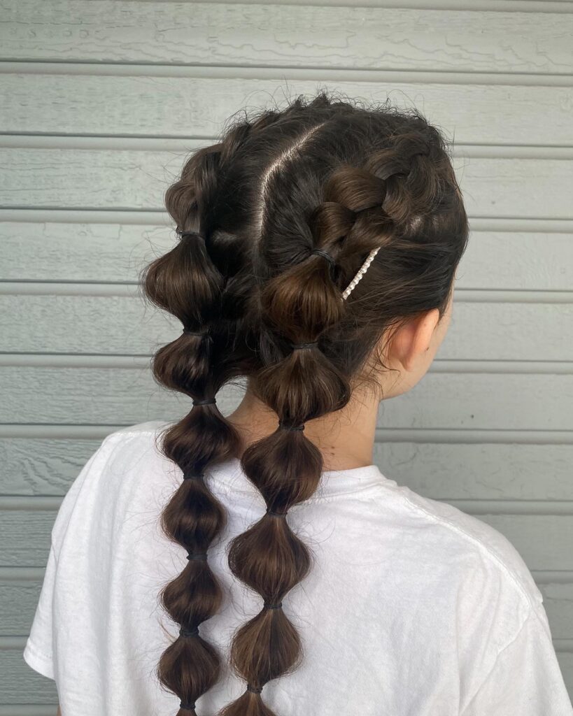 A woman from the back shows dark brown Dutch braids that flow into voluminous bubble pigtails accented with a pearl hair clip.