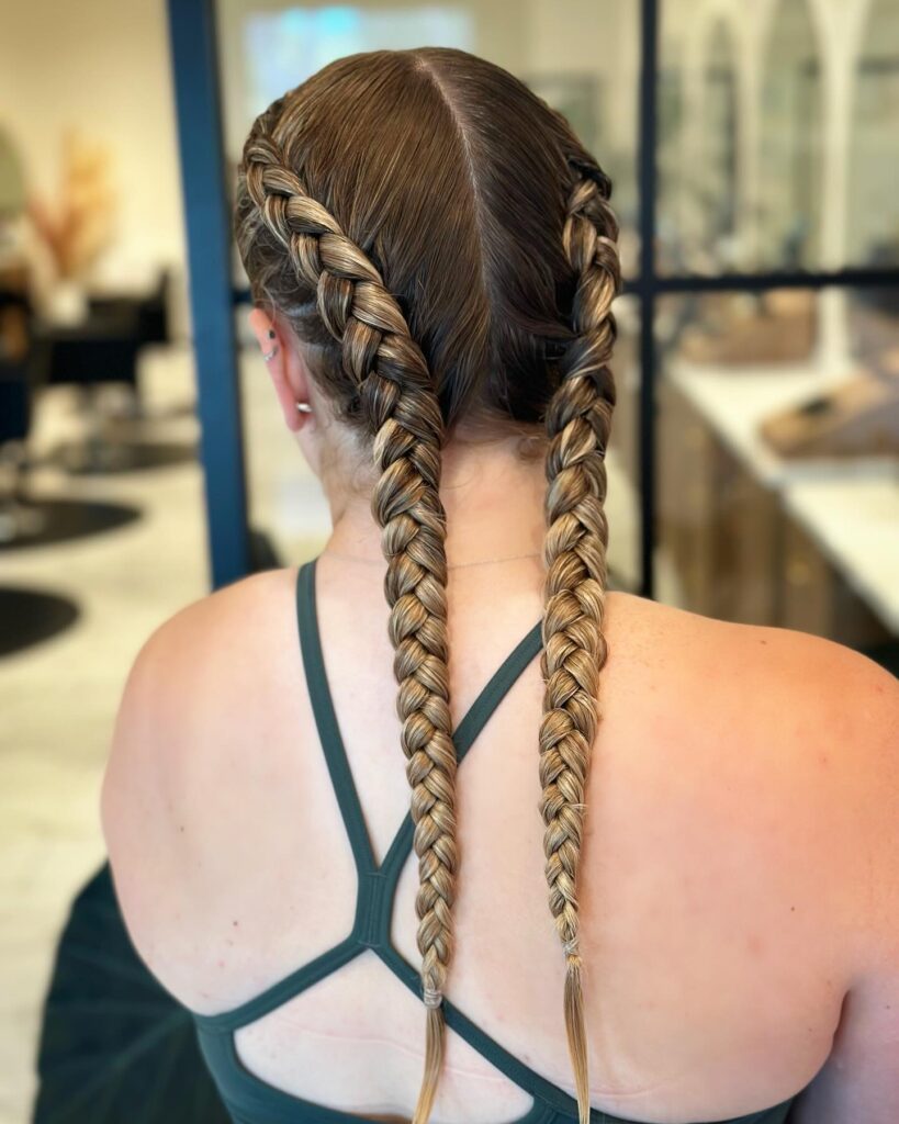 A view from behind of a woman with long and sleek double Dutch braids featuring caramel highlights and brunette roots.