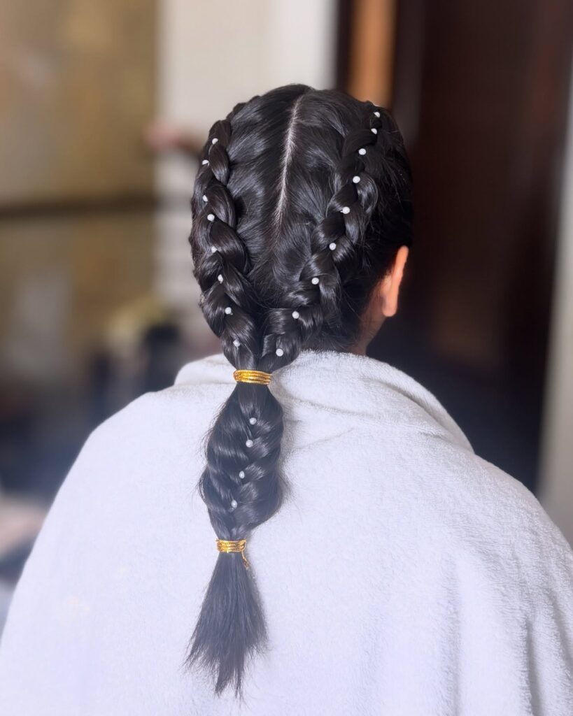 A view from behind of a woman with jet black Dutch braids merging into one single plait decorated with small white pearls and gold hair ties.