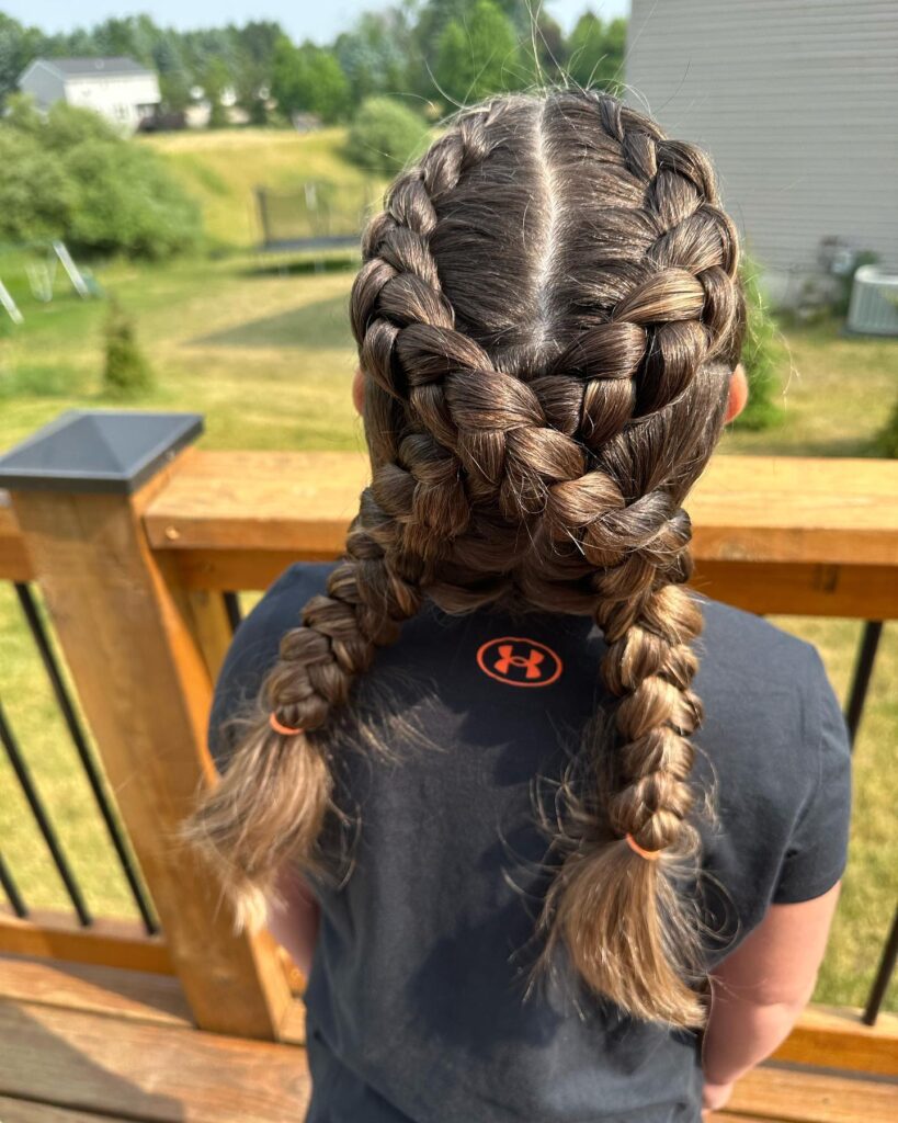 A view from behind of a girl wearing thick brunette double Dutch braids that criss-cross at the nape of the neck and are secured with orange elastics.