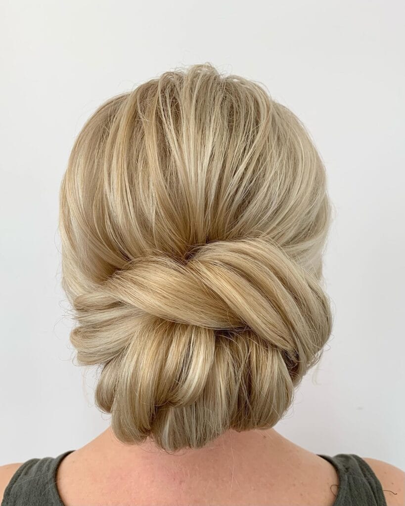 A back view of a woman with highlighted blonde hair styled in a sophisticated low chignon bun with an elegant overlapping wrap detail.
