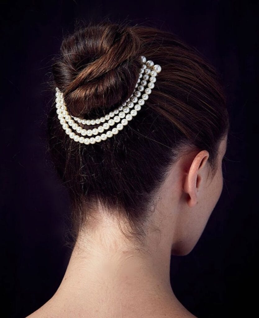 A back view of a woman with dark brown hair in a sleek chignon bun decorated with three cascading strands of white pearls.