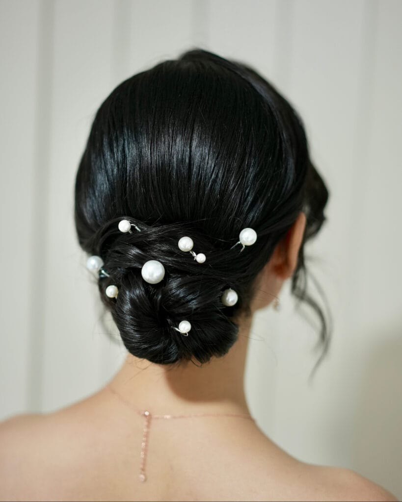 A back view of a woman with sleek black hair styled in a twisted low chignon bun decorated with various-sized white pearl pins.