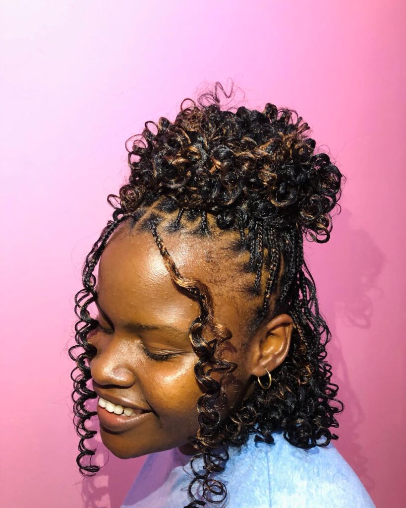 A smiling woman wearing a half-up hairstyle with small braids and voluminous copper-highlighted curls on top.