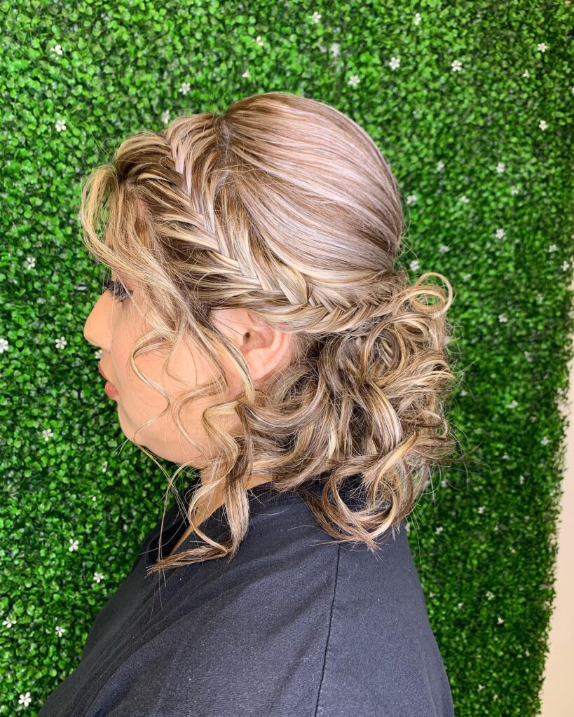 A side profile of a woman with highlighted blonde hair styled in a thick side braid and a messy curly low bun.