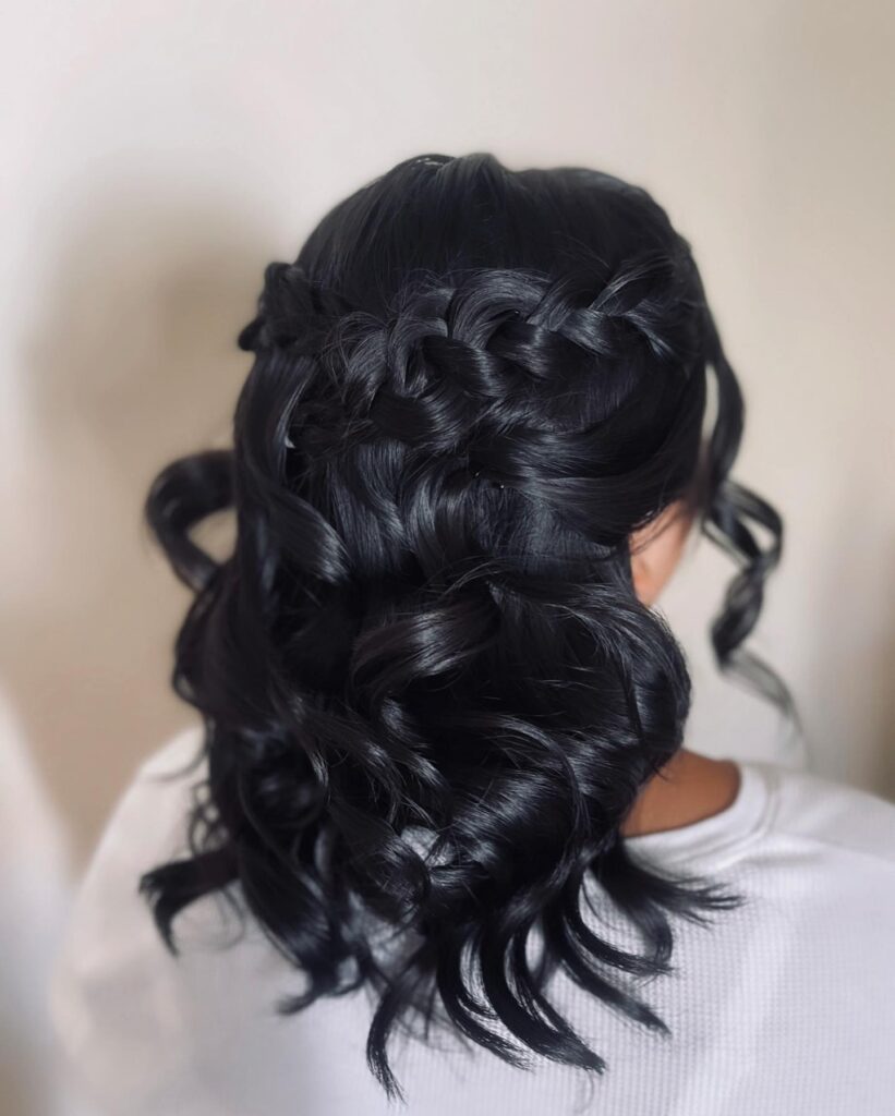 A rear view of a woman with shiny black hair styled in a thick half-up braid and large soft curls.