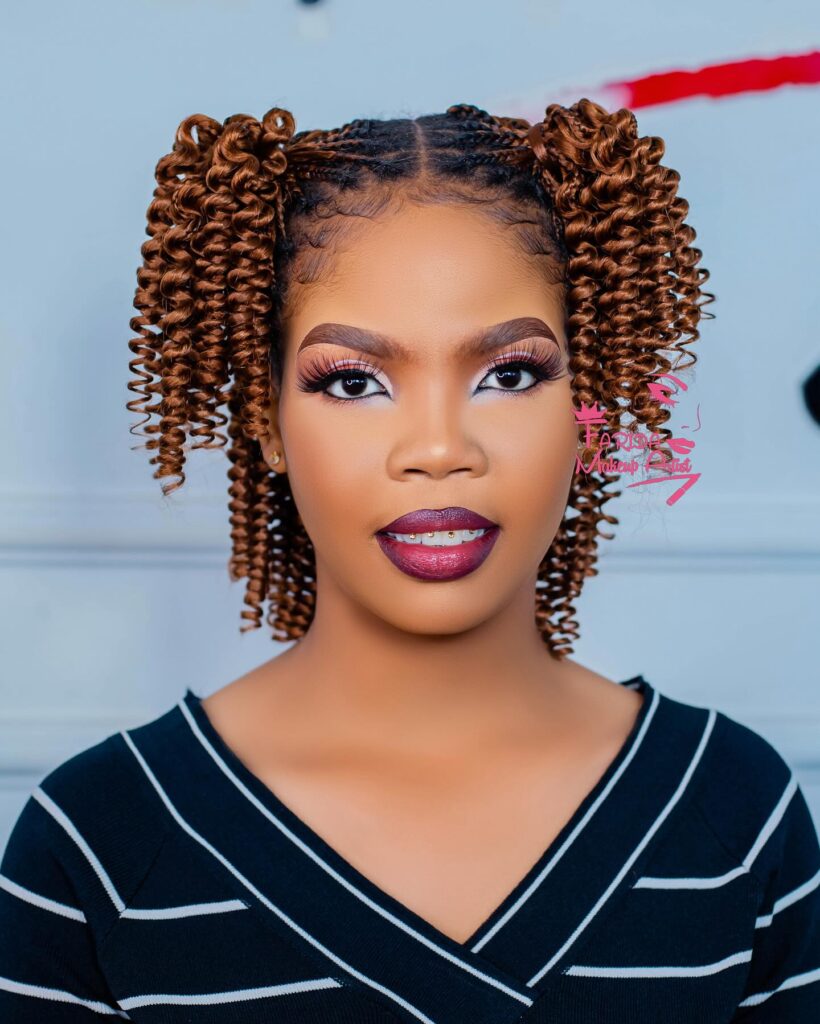 A woman with auburn hair styled in two high curly pigtails with small braids at the roots.