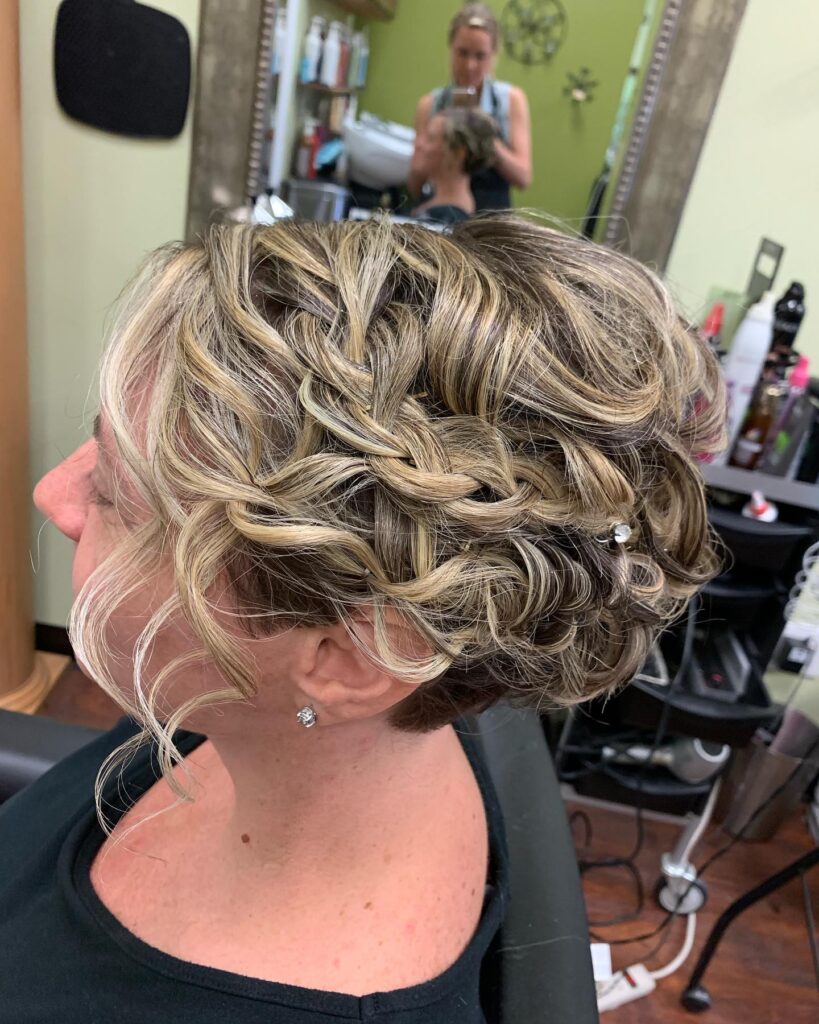 A side view of a woman with highlighted ash blonde hair styled in a braided updo with soft curls and a small jewel hairpin.