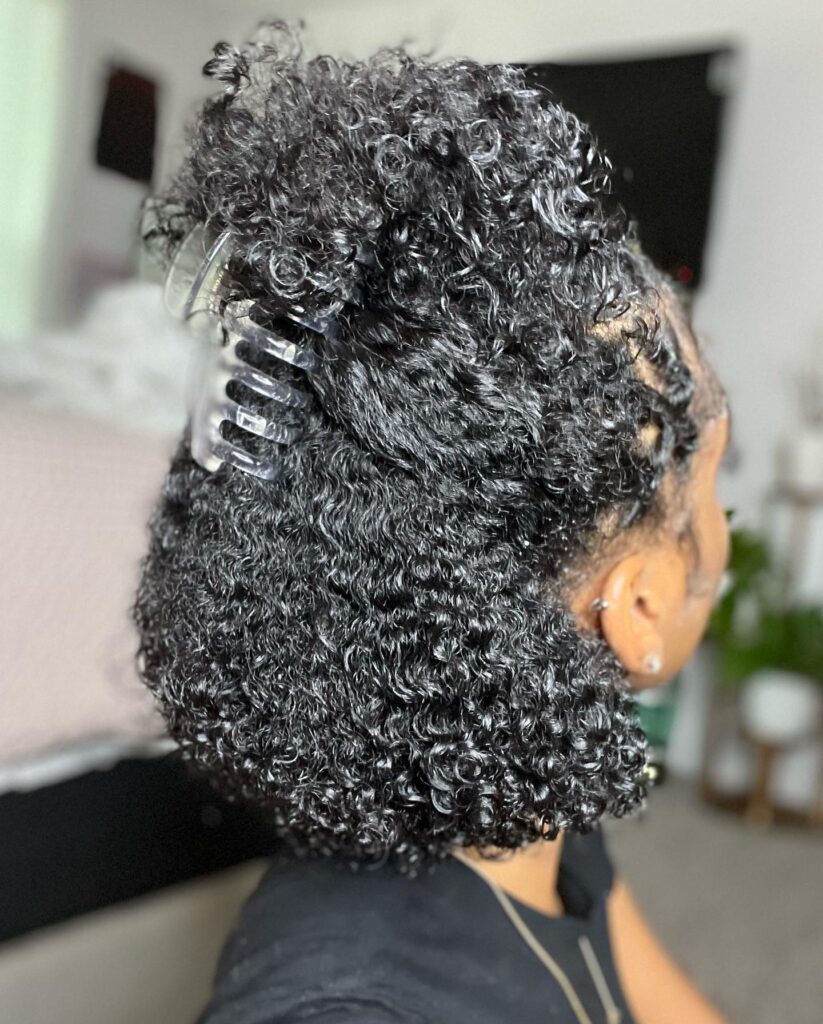 A woman with short natural black curls styled in a half-up look secured by a clear claw clip at the crown.