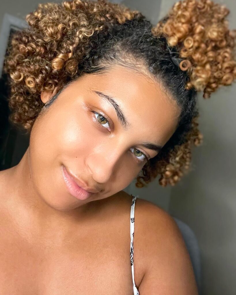 A woman with short natural curls styled into two high space puffs with honey blonde highlights.