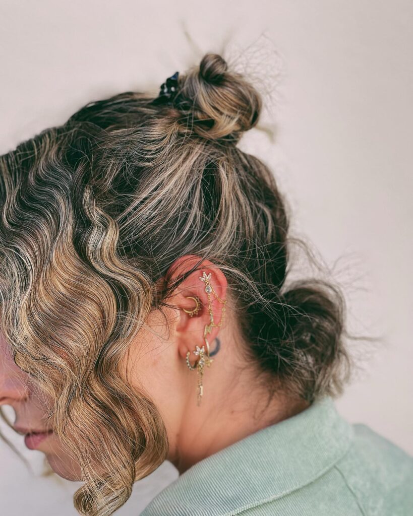 A side view of a woman with short, wavy blonde hair styled in a messy half-up topknot.