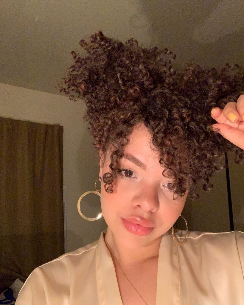 A woman with tight dark brown curls styled in a high messy bun with curly bangs.