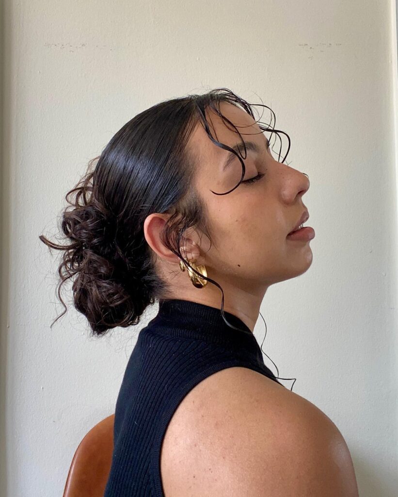 A side profile of a woman with dark hair styled in a sleek, low bun with wet-look curly tendrils.