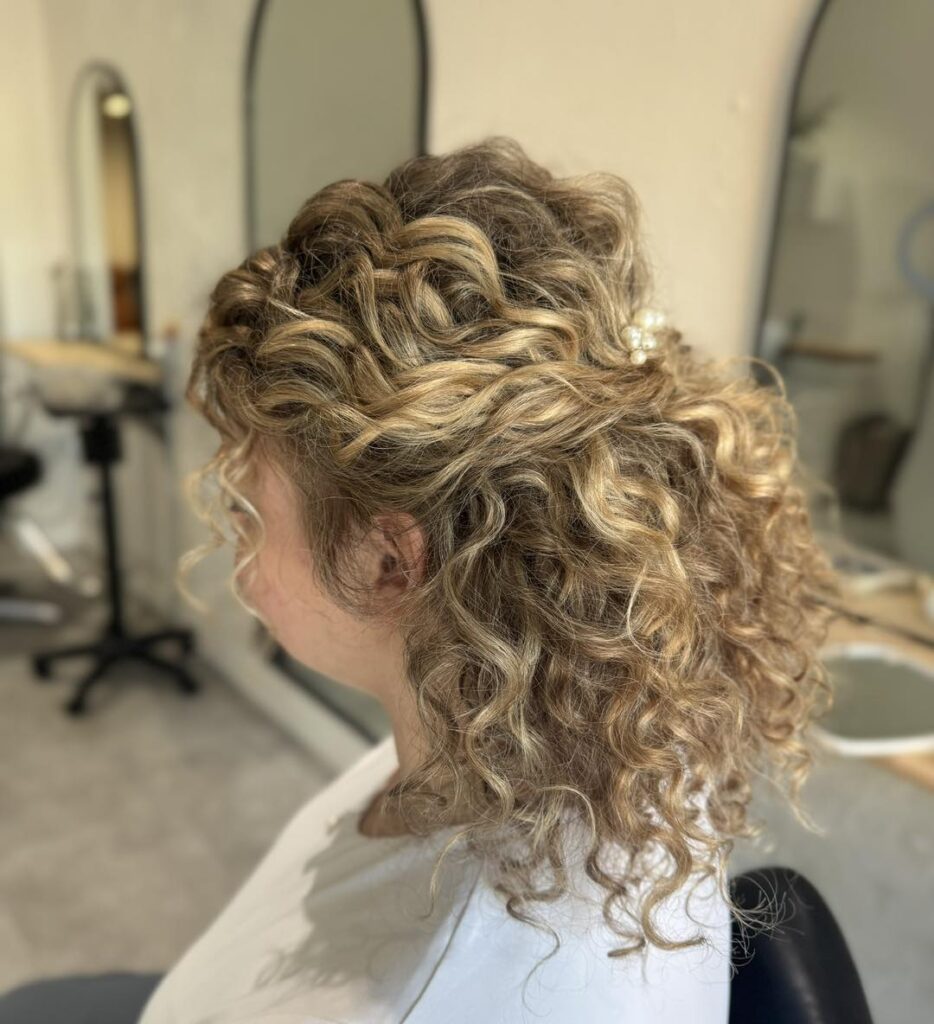 A side view of a woman with medium-length, honey-blonde curly hair styled in a half-up look with pearl hairpins.