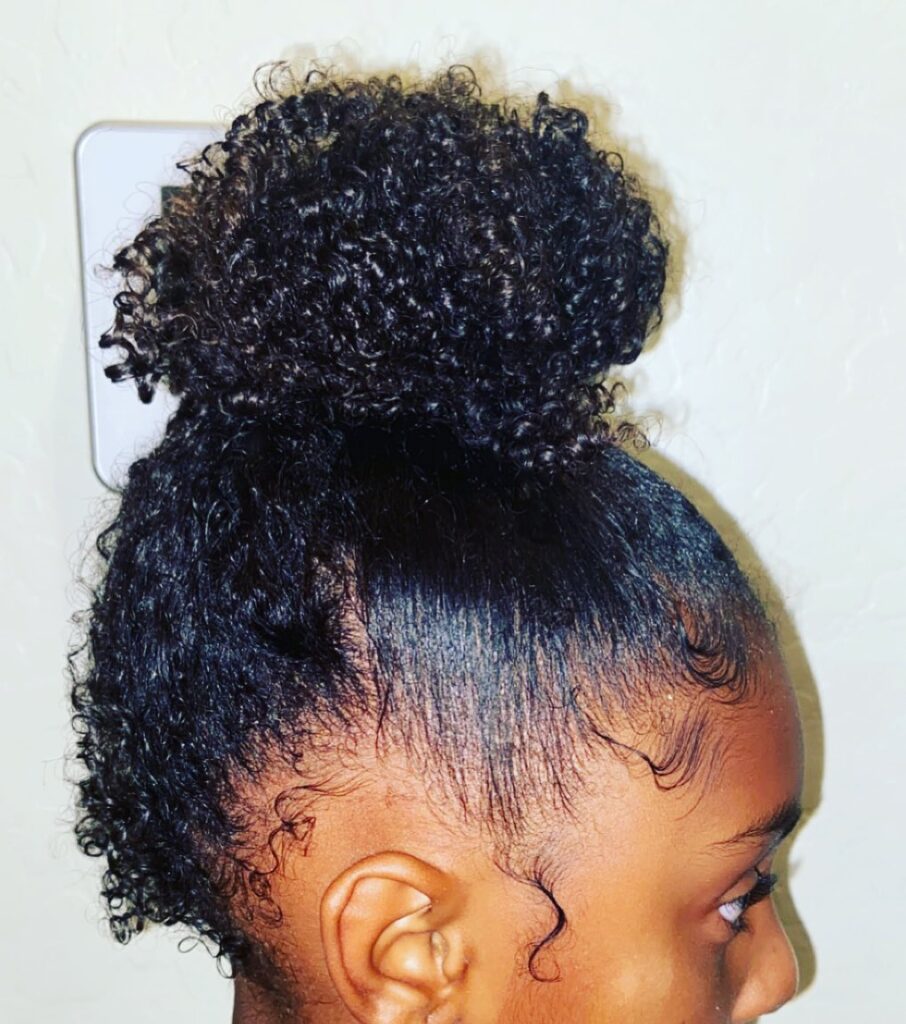 A side profile of a person with natural black curls styled in a high top knot with detailed baby hairs along the hairline.