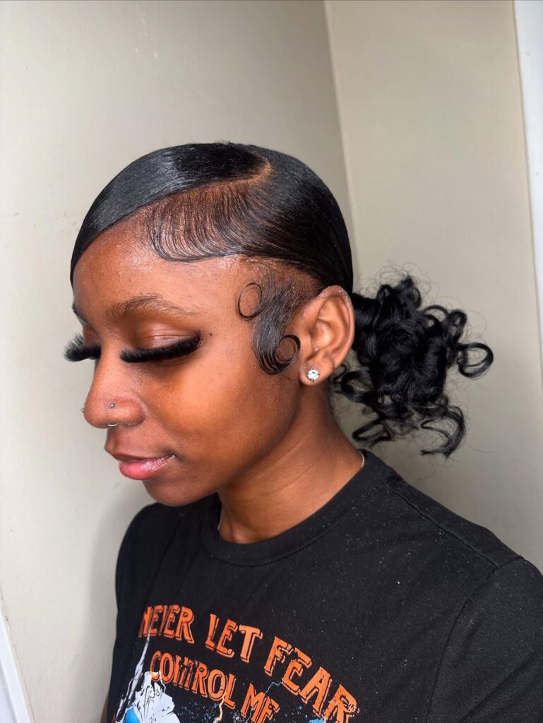 A side profile of a woman with black hair styled into a sleek, low, curly ponytail with intricate laid edges.