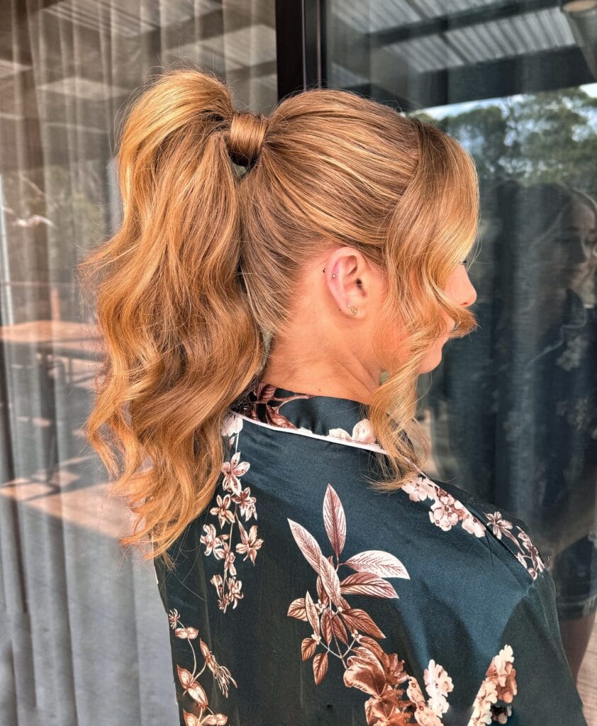 A woman from the side is showing a high honey-blonde ponytail with soft waves and face-framing strands.
