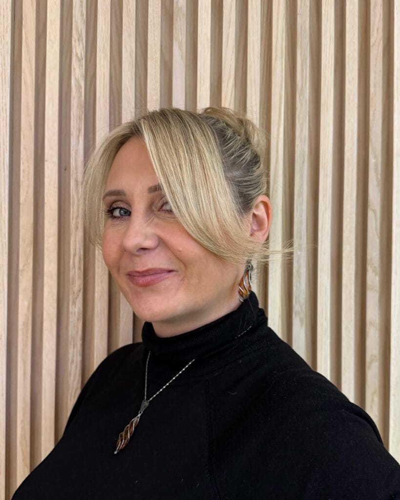 A blonde woman wearing a black turtleneck with her hair in a neat updo and long side-swept bangs.