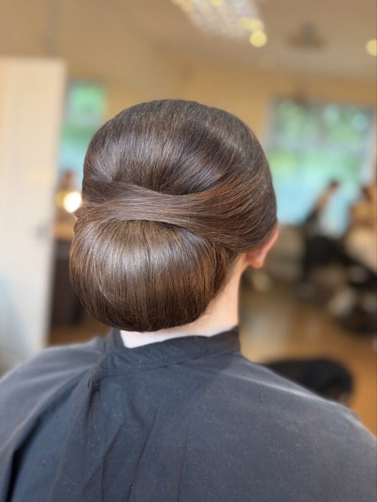 A back view of a woman with rich brunette hair styled into a smooth and voluminous low chignon with a crossover wrap detail.