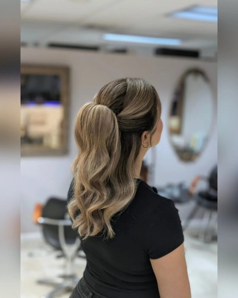 A back view of a woman with ash-blonde wavy hair styled into a mid-height ponytail with twisted side details.