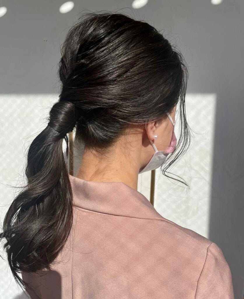 A back view of a woman with dark wavy hair in a low ponytail with a hair-wrapped base and loose side strands.