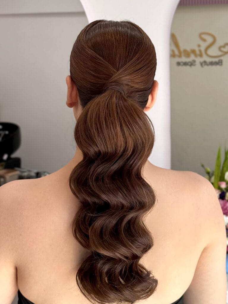 A back view of a woman with long brunette hair styled into a sleek ponytail with deep, structured Hollywood waves.