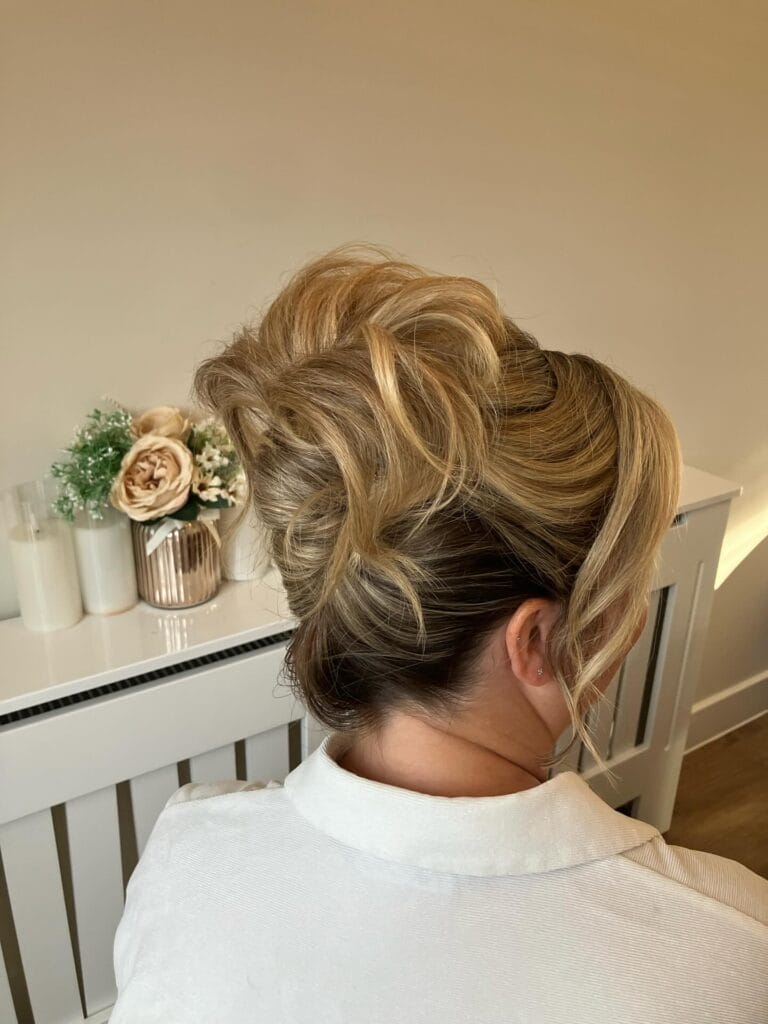 A back view of a woman with highlighted blonde hair styled in a voluminous French pleat updo with loose side curls.