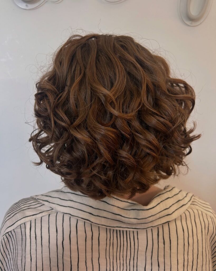 A back view of a woman with neck-length layered curly hair in a rich, glossy chestnut brown shade.