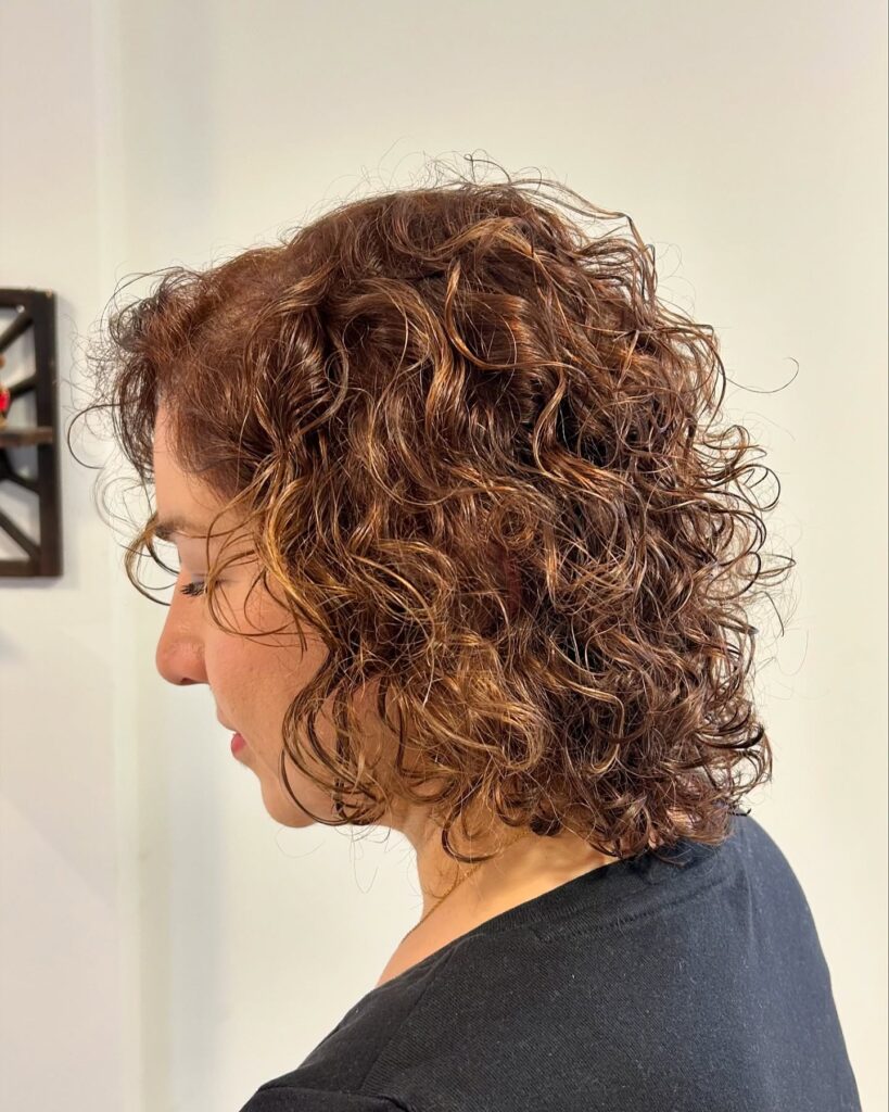 A profile view of a woman with a shoulder-length curly lob featuring auburn and caramel balayage highlights.