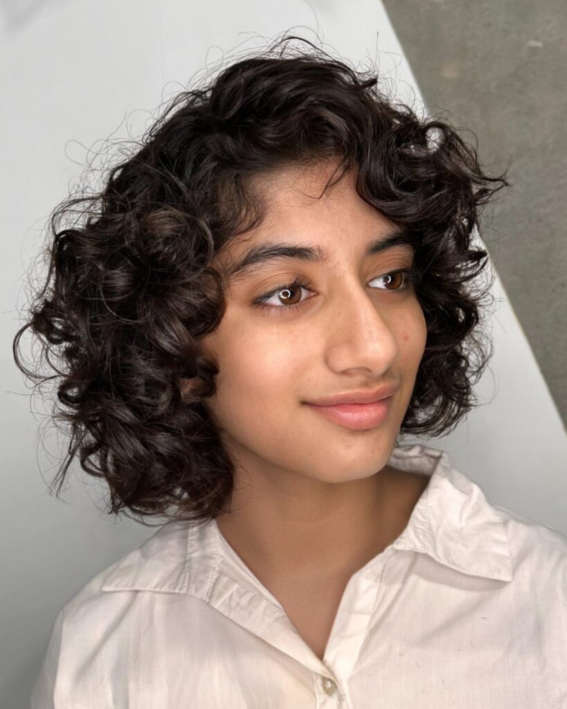 A young woman with a chin-length dark espresso curly bob featuring voluminous layers and a deep side part.