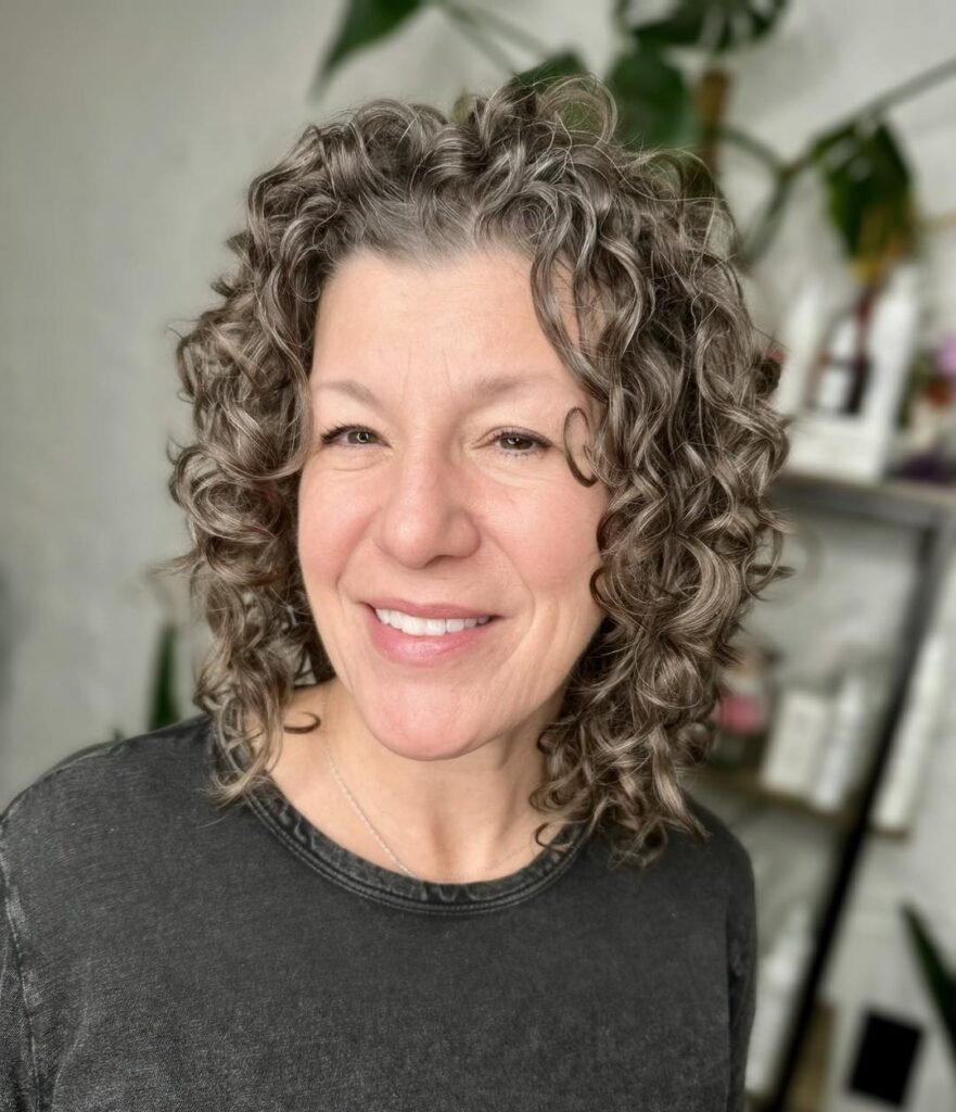 A smiling woman with a shoulder-length salt-and-pepper curly lob featuring defined spirals and voluminous layers.