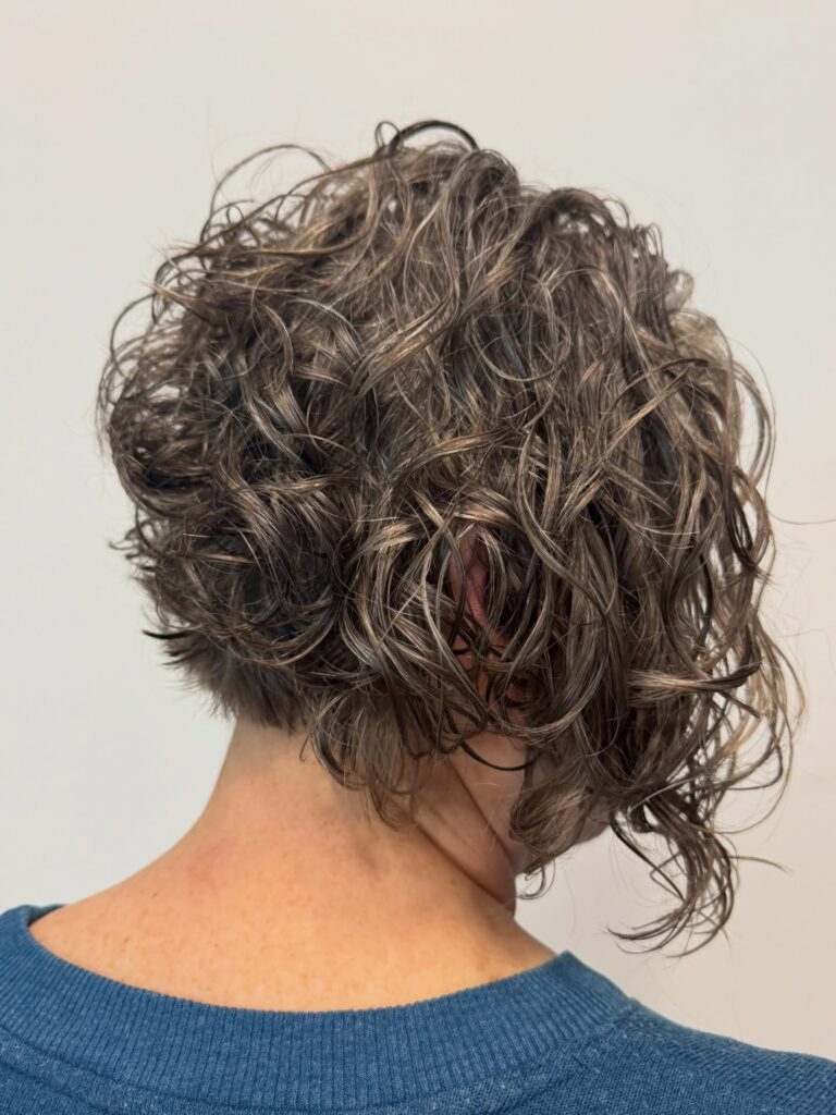 A back view of a woman with a short, stacked curly bob in a cool ash brown shade featuring a tapered nape.