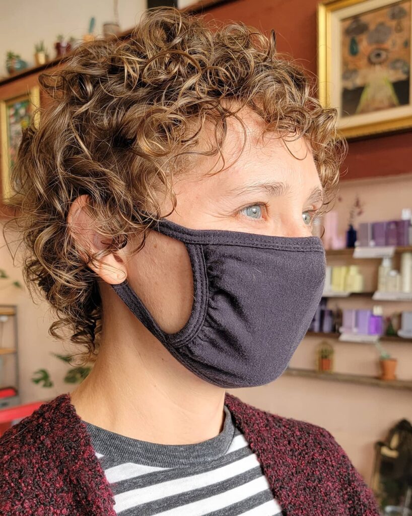 A close-up shot of a woman with a curly, honey-brown cropped shaggy hairstyle wearing a dark face mask.