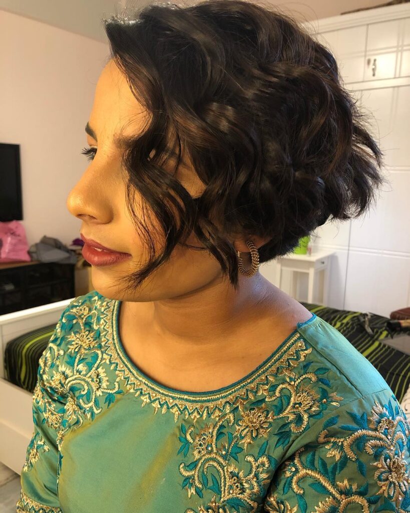 A woman with voluminous short brunette curls styled in a deep side part and wearing a teal embroidered top.