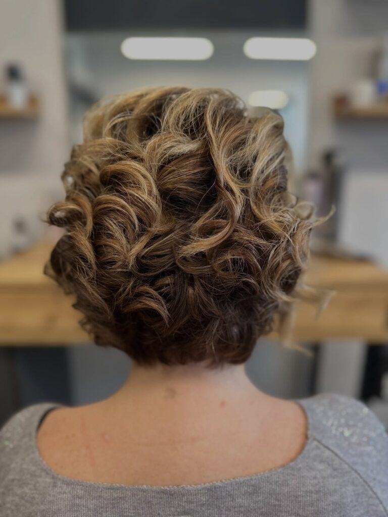 A back view of a woman with a stacked curly bob featuring blonde and brown highlights.